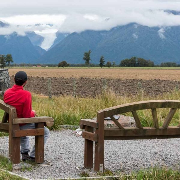Homem sentado em banco observa vista do glaciar Fox