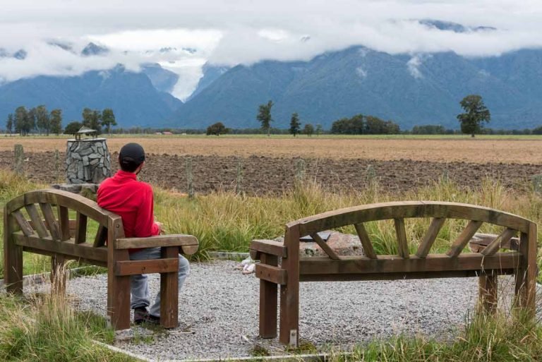 Homem sentado em banco observa vista do glaciar Fox